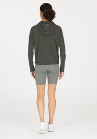 Dunkelgrüne Kapuzenjacke mit langen Ärmeln, getragen über grauen Fahrradhosen. Der Stoff wirkt glatt und das Design ist figurbetont und minimalistisch.