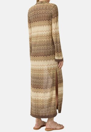 Femme portant une longue robe tricotée à motif chevron dans des tons marron et beige, avec une fente sur le côté, associée à des sandales plates à brides.