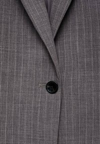 Nahaufnahme eines grauen Nadelstreifen-Blazers mit Kerbkragen und einem einzelnen schwarzen Knopf, der geschlossen ist.