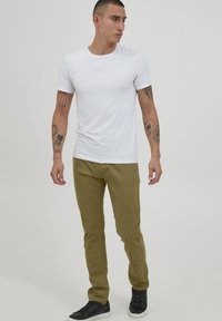 T-shirt blanc à manches courtes associé à un pantalon slim vert olive et des baskets noires. Présente des tatouages sur les bras et une expression neutre.