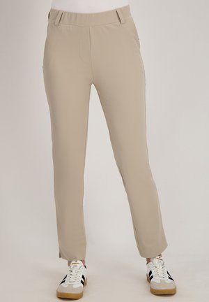 Beige slim-fit broek met een elastische tailleband, gedragen met witte en zwarte vetersneakers tegen een effen achtergrond.