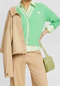 Grün gestrickter Cardigan mit weißen Knöpfen, darüber getragen ein grün-weiß gestreiftes Hemd. Beige Jacke und grüne Handtasche vervollständigen das Outfit.