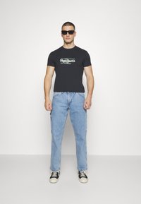 Camiseta de algodón negra con el logo "Pepe Jeans London", combinada con pantalones cargo de mezclilla en azul claro. Accesorios incluyen gafas de sol negras y zapatillas.