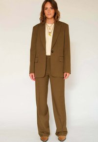 Costume marron sur mesure avec un blazer à un bouton et un pantalon droit. Comprend des poches latérales et des colliers en or superposés.