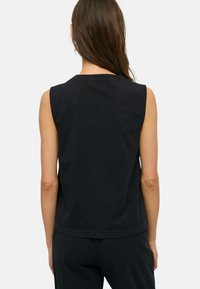 Mujer con el cabello largo y castaño, con una camiseta negra sin mangas y pantalones negros, de espaldas contra un fondo claro y sencillo.