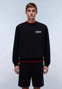 Ung mann iført en svart hettegenser med logo på brystet og svarte shorts, står mot en ensfarget lys grå bakgrunn.