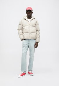 Pufová bunda vo svetlo béžovej farbe, so zipsovým zapínaním a horizontálnym prešívaním. V kombinácii so svetlo modrými džínami a červenými teniskami.