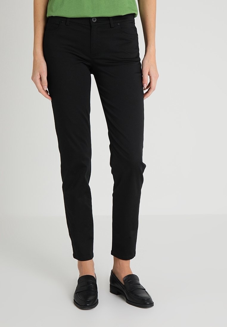 Femme portant un pantalon noir slim et des mocassins noirs, debout devant un fond clair uni.