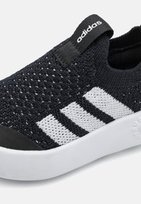 Sorte strikkede slip-on sko med hvit tre-stripes design. Har teksturert overdel, svart gummitå og hvit såle med subtil merkevare.