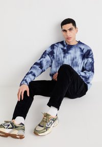 Modra in bela tie-dye pulover, črne kordne hlače in večbarvni športni čevlji, ki ležijo na beli površini. Priložnostna obleka z kontrastom tekstur.