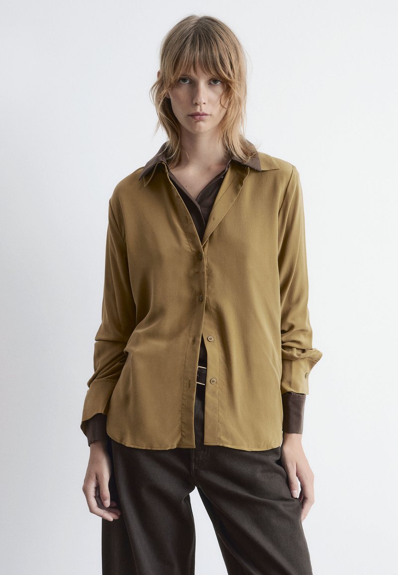Brown long-sleeve shirt with a button front and a contrasting collar. Soft fabric with a relaxed fit and slightly rounded hem.