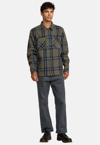 Camicia di flanella a quadri in verde oliva con linee blu e nere, dotata di due tasche sul petto e chiusura a bottoni; abbinata a pantaloni grigi e scarpe nere.