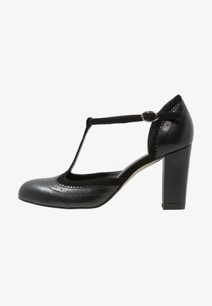 Scarpa nera in pelle con tacco alto, cinturino a T, dettagli ondulati e forature; il tacco spesso offre stabilità e supporto.