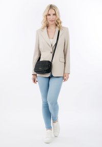 Frau geht vorwärts, trägt beige Blazer, hellblaue Jeans, weiße Turnschuhe und eine schwarze gesteppte Umhängetasche vor weißem Hintergrund.