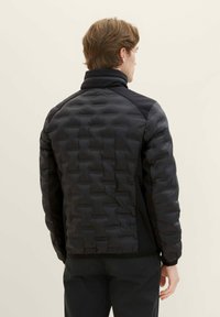 Giacca a vento nera con design trapuntato e collo alto. I pannelli laterali in un materiale nero liscio aumentano la flessibilità e la vestibilità.
