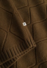 Smeđa pletena deka s geometrijskim uzorkom, textured površinom i rebrastim rubovima. Ima malu bijelu oznaku s slovom "W."