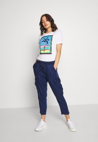 Camiseta blanca con un gráfico colorido de una palmera. Pantalones cargo azul marino con bolsillos, cintura ajustable con cordón y puños acanalados. Zapatillas blancas.
