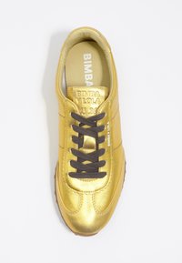 Złote metaliczne sneakersy o gładkiej fakturze, czarne sznurowadła i branding na języku. Posiadają zaokrąglony kształt oraz jasnobrązową podeszwę.