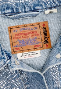 Hele helesinine teksajakk, millel on pärlisemed, tan värvi nahklabel sinise tekstiga ja allpool valge kangasetikett, millel on märgistus "LEVI'S."