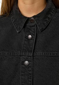 Svart denimjakke med spiss krage, metallknapper og teksturert stoff. Designet har synlige sømmer langs sømmene.