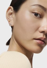 Dos pendientes: una estrella de oro y un diseño circular plateado con pequeñas piedras claras. Textura sutil de la piel y una blusa de punto crema visible.