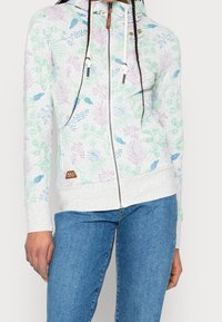 Ljusgrå zip-up hoodie med blommönster i grönt, blått och lila. Har dragsko runt huven och subtil logotyp-patch på fållen. Mjuk tyg.