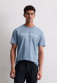 Lichtblauwe katoenen t-shirt met korte mouwen, voorzien van witte tekstlogo en boodschap gecentreerd op de voorkant. Draagt bij donkere broek.