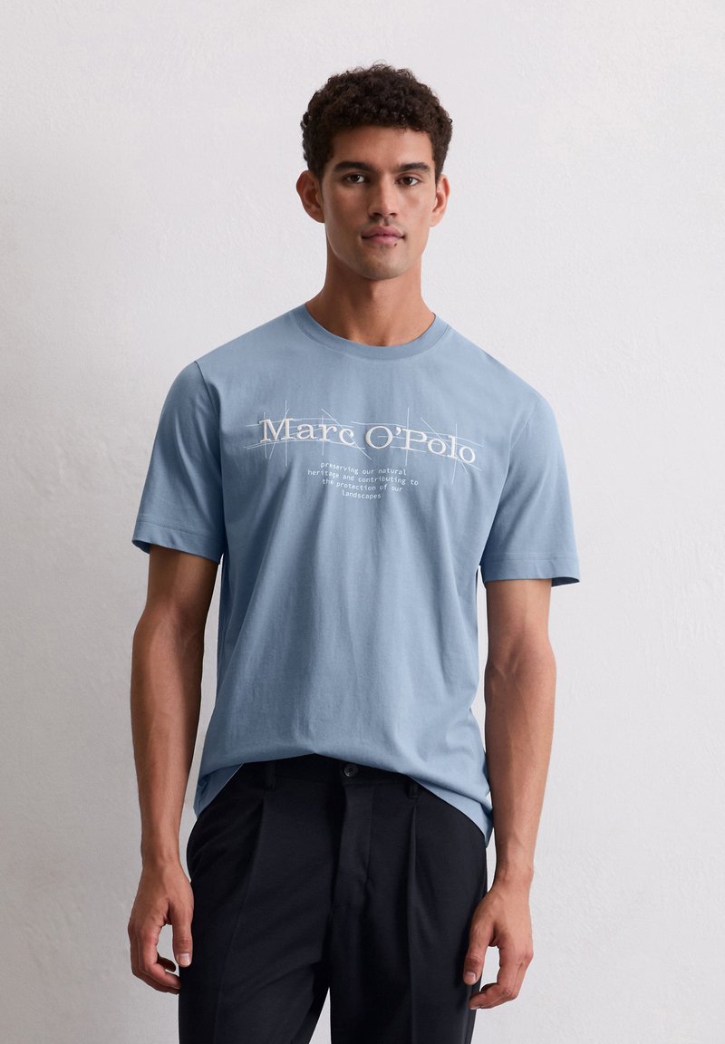 Lichtblauwe katoenen t-shirt met korte mouwen, voorzien van witte tekstlogo en boodschap gecentreerd op de voorkant. Draagt bij donkere broek.