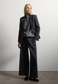 Blazer grigio scuro su un dolcevita grigio, abbinato a pantaloni larghi e scarpe nere, indossato da un modello su uno sfondo neutro.
