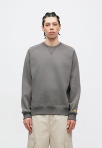 Gray crewneck sweatshirt with raglan sleeves, ribbed cuffs, and hem. Features a small yellow logo on the left sleeve. Smooth texture.