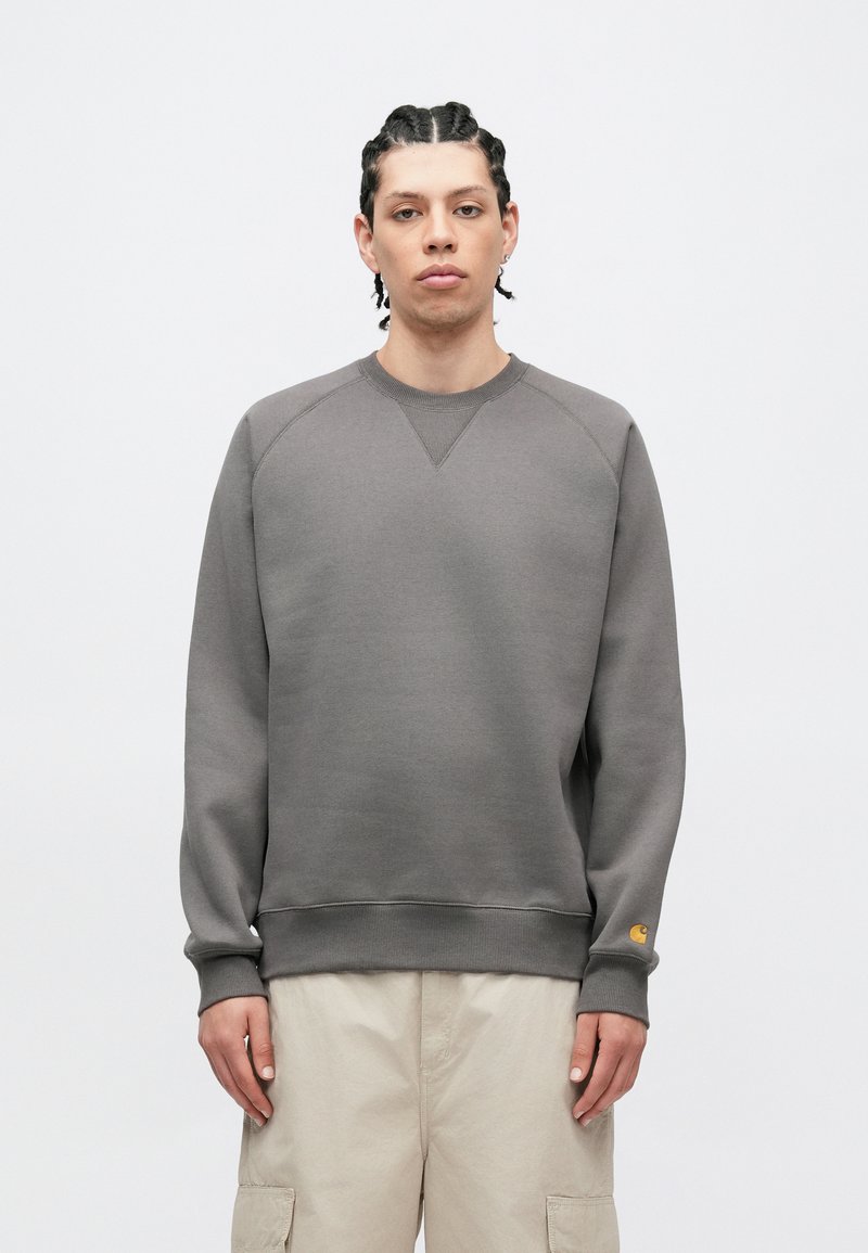 Grå crewneck sweatshirting med raglanærmer, ribbet manchetter og kant. Har en lille gul logo på venstre ærme. Glat tekstur.