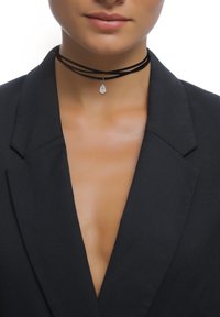 Schwarzes Wildleder-Choker mit mehreren Strängen, das einen hängenden, tropfenförmigen silbernen Anhänger zeigt. Das Modell trägt einen Blazer mit tiefem V-Ausschnitt.