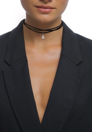 Schwarzes Wildleder-Choker mit mehreren Strängen, das einen hängenden, tropfenförmigen silbernen Anhänger zeigt. Das Modell trägt einen Blazer mit tiefem V-Ausschnitt.
