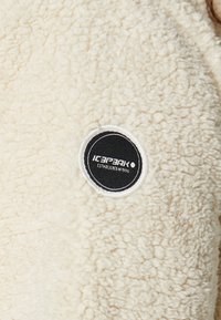 Tela de forro polar color crema con una superficie suave y texturizada. Presenta una etiqueta negra redonda con "ICEPEAK ESTABLECIDO EN 1996" en texto blanco.