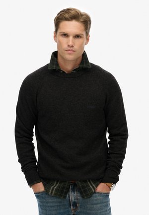 Suéter gris oscuro con un escote redondo, usado sobre una camisa de cuadros verdes. El suéter tiene puños acanalados y un corte relajado. Se ven jeans de mezclilla.