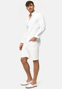 Witte linnen shirt met een kraag, lange mouwen en knoopdetails, gecombineerd met witte shorts en witte espadrilles, tegen een effen achtergrond.