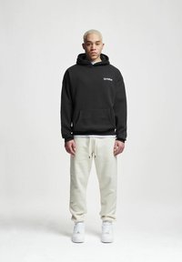 Must värviga kapuutsiga dressipluus, millel on ees tasku ja valge logo. Kombineeritud vabade, heleda beeži värvi dressipükstega ja valgete tossudega. Lihtne, juhuslik riietus.