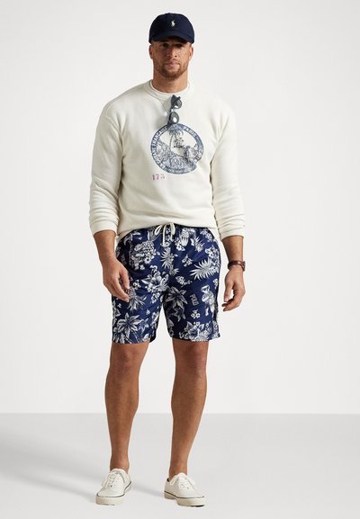 Balta džemperis ar grafisku apdruku, tumši zili ziedu pludmales šorti un balti sporta apavi. Aksesuāri ietver saulesbrilles un tumši zilu cepuri.