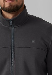 Giacca grigia con zip realizzata in materiale morbido. Presenta un alto colletto, cuciture sottili e un piccolo logo sul petto.