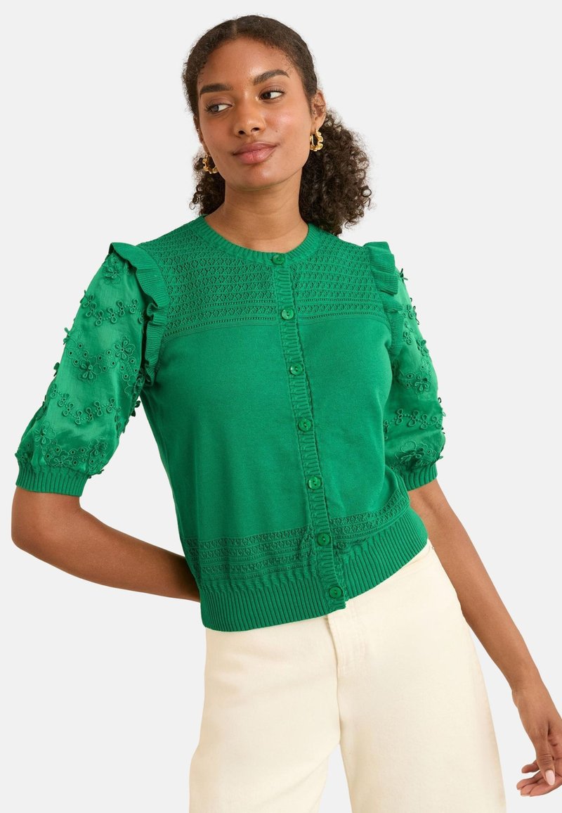 Femme aux cheveux bouclés portant un cardigan vert texturé boutonné avec des manches bouffantes brodées de fleurs et un pantalon crème sur fond blanc.