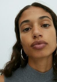 Close-up of a person with medium brown skin, long dark hair, wearing large silver hoop earrings and a gray high-neck top. Lips are slightly parted.