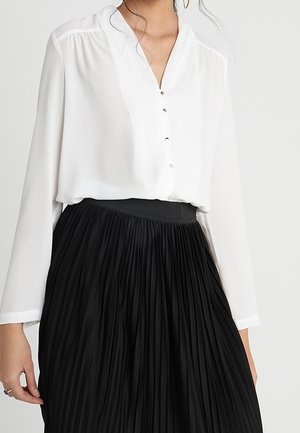 Vrouw met een witte blouse met lange mouwen en zilveren knopen, ingestopt in een zwart geplooide hoge taille rok, uitgeknipt ter hoogte van het midden van de torso.