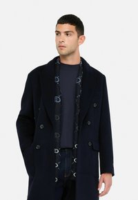 Palton din lână bleumarin cu design dublu, revere mari, două buzunare frontale și o eșarfă cu model care include accente de albastru închis.