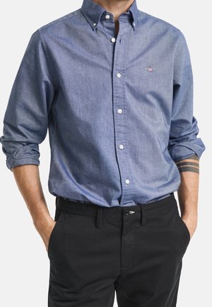 Mann trägt ein blaues Hemd mit Knöpfen, die Ärmel hochgekrempelt, und schwarze Hose, die Hände locker in den Taschen, ein Arm-Tattoo sichtbar.