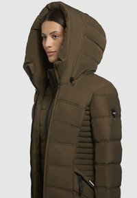 Bruine pufferjack met een grote capuchon, horizontaal geborduurd design, met rits afsluitbare zakken en een logo-accent op de mouw. Soepele textuur.