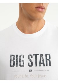 Közeli kép egy személyről, aki fehér pólót visel, melyen feketével a "BIG STAR" felirat és egy kis csillag ikon van nyomtatva a mellrészen.