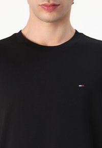 Schwarzes Baumwoll-T-Shirt mit rundem Ausschnitt und kurzen Ärmeln, mit einem kleinen, mehrfarbigen Logo auf der linken Brustseite.