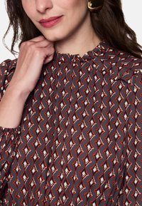 Graue Bluse mit geometrischem Rautenmuster in Rot und Creme. Mit hohem Kragen und gerafften Ärmeln.