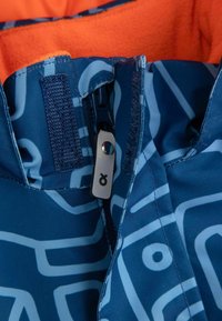 Niebieska kurtka narciarska z zamkiem, ozdobiona odważnym, abstrakcyjnym wzorem w jaśniejszym odcieniu niebieskiego. Wewnątrz wyłożona jasnopomarańczowym polarem dla zapewnienia ciepła.