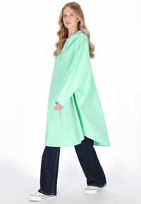 Mentaszínű, könnyű esőkabát kapucnival, elülső zsebbel és laza szabással, sötét farmer bőszárú nadrággal és fehér tornacipővel párosítva.